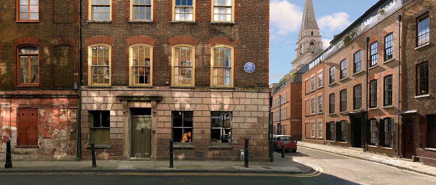 Photograph showing corner of brick terrace with blue plaque mounted at first floor level and a church spire visible in distance